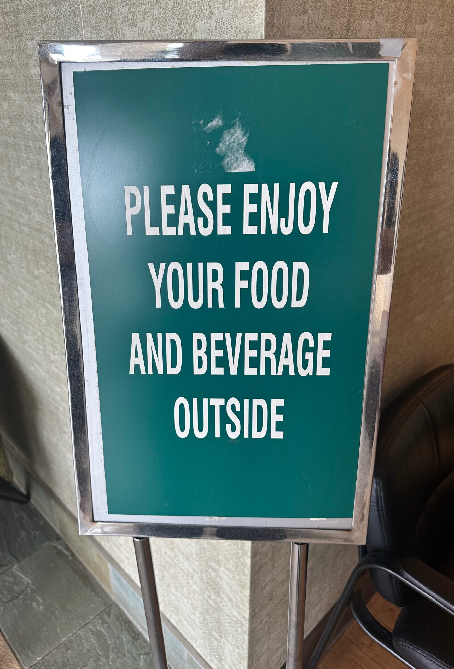 A framed indoor sign in a chrome stand displaying white block text on a green background that reads: "PLEASE ENJOY YOUR FOOD AND BEVERAGE OUTSIDE."